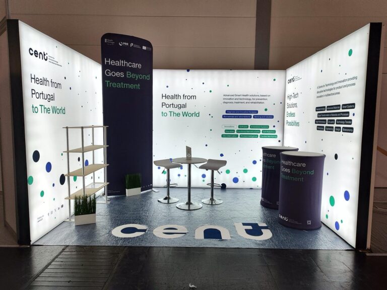 Stand da Centi finalizado, com um design moderno e iluminado. O espaço promove soluções tecnológicas para a saúde, com frases como "Healthcare Goes Beyond Treatment" e elementos gráficos em tons de azul, verde e branco. Inclui mesas altas, prateleiras e pequenos detalhes decorativos.